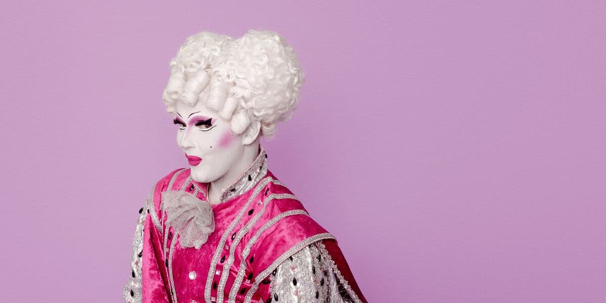 The image shows a performer dressed in an elaborate pink and silver 18th-century–style costume, complete with a powdered wig, white stockings, and dramatic stage makeup, posing joyfully against a pastel purple background.