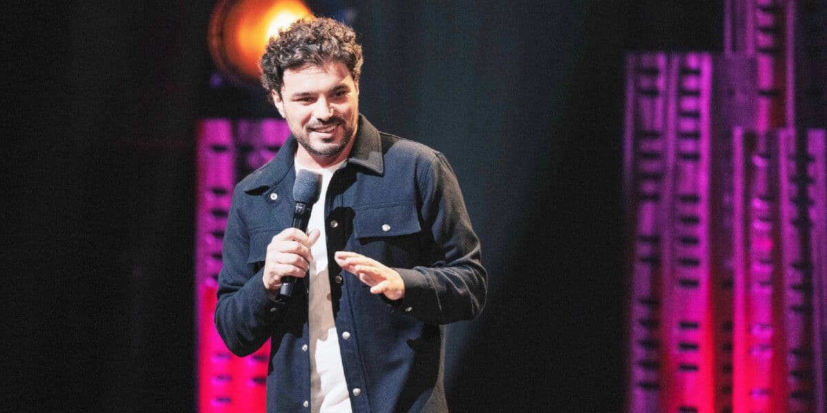 Amos Gill is onstage wearing a dark denim jacket & white tshirt and holding a microphone in his right hand as he gestures downwards with left palm. He has with short, thick dark hair, dark eyes and facial stubble and is smiling broadly as the audience is clearly eating up his story.