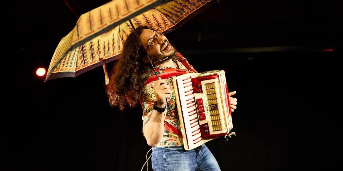 Accordion Ryan holds an accordion, wearing a flowered shirt and jeans. He plays the accordion with one hand, while holding an umbrella over his head with the other.